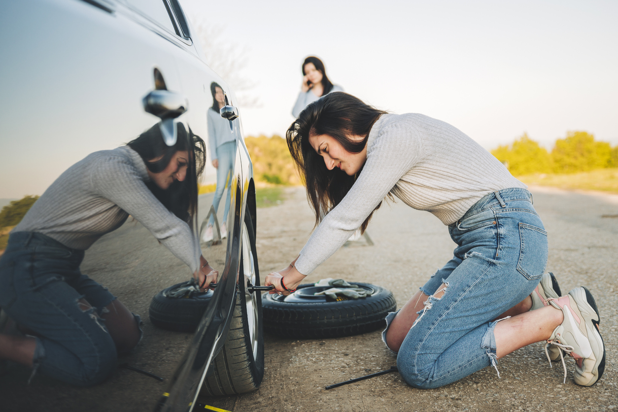 Woman changing a flat tire on the side of the road while her friend calls Savage L&B for a tow to their service center