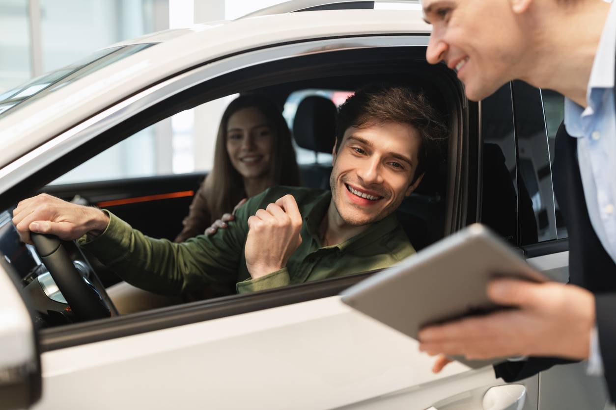 Young Caucasian couple clarifying car purchase details with salesman, looking at tablet screen in dealership center