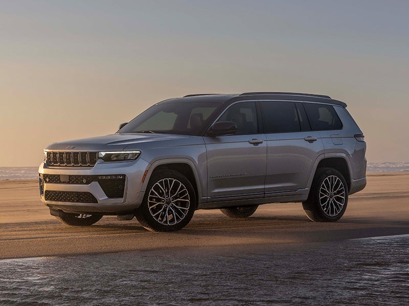 A silver SUV is parked on a sandy beach at sunset, with the ocean and sky visible in the background.