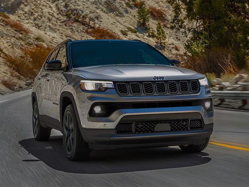 A silver Jeep SUV drives on a paved mountain road, with rocky terrain and trees visible in the background.