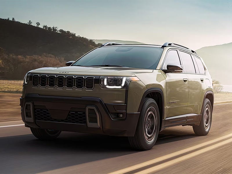 A light green Jeep SUV drives along a road through hilly terrain at sunrise or sunset.
